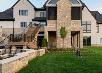 Modern two-story home with brick chimney, stone facade, and outdoor staircase; green lawn and a dog in the yard.